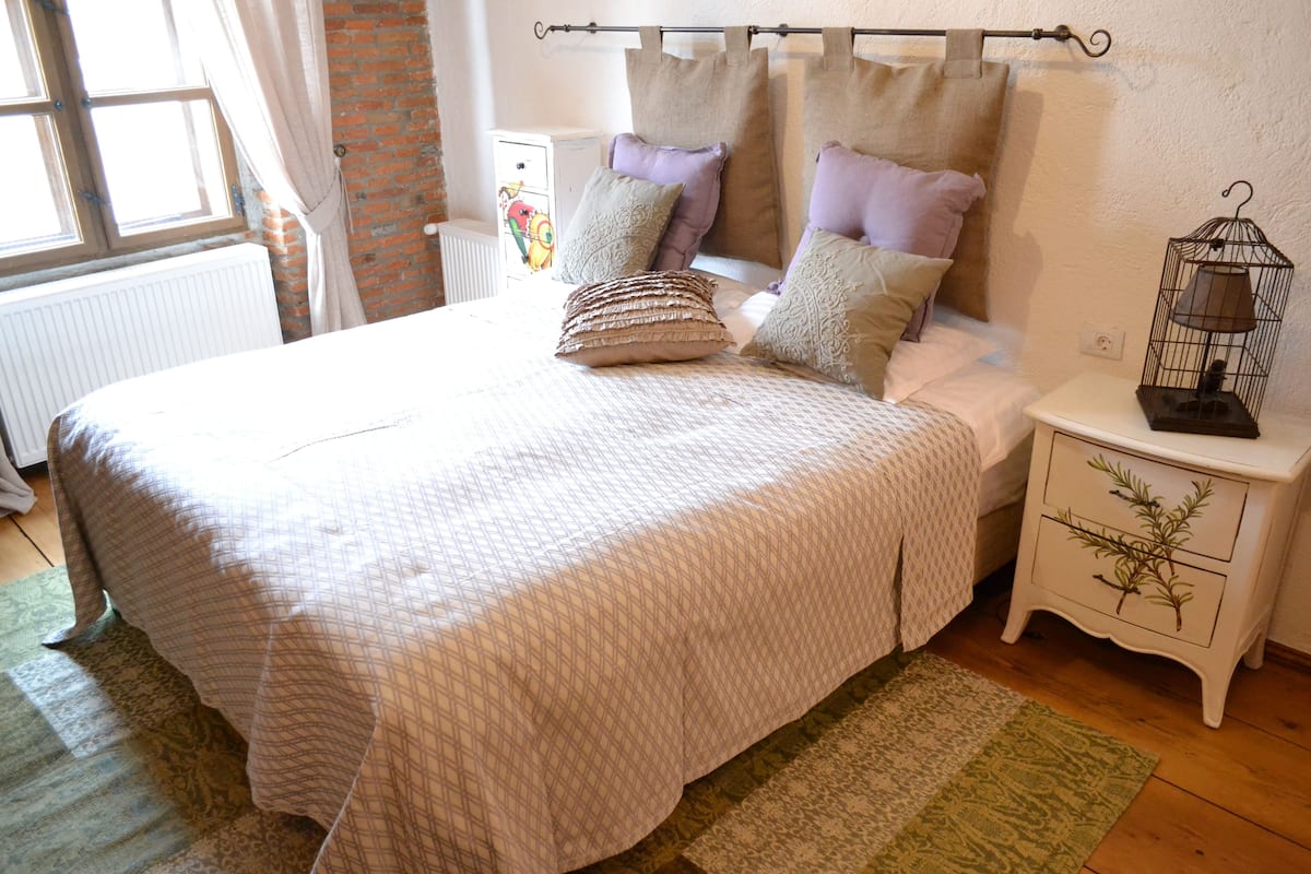 A neatly made bed with a patterned bedspread, a bedside table with a lamp, and a window with curtains.