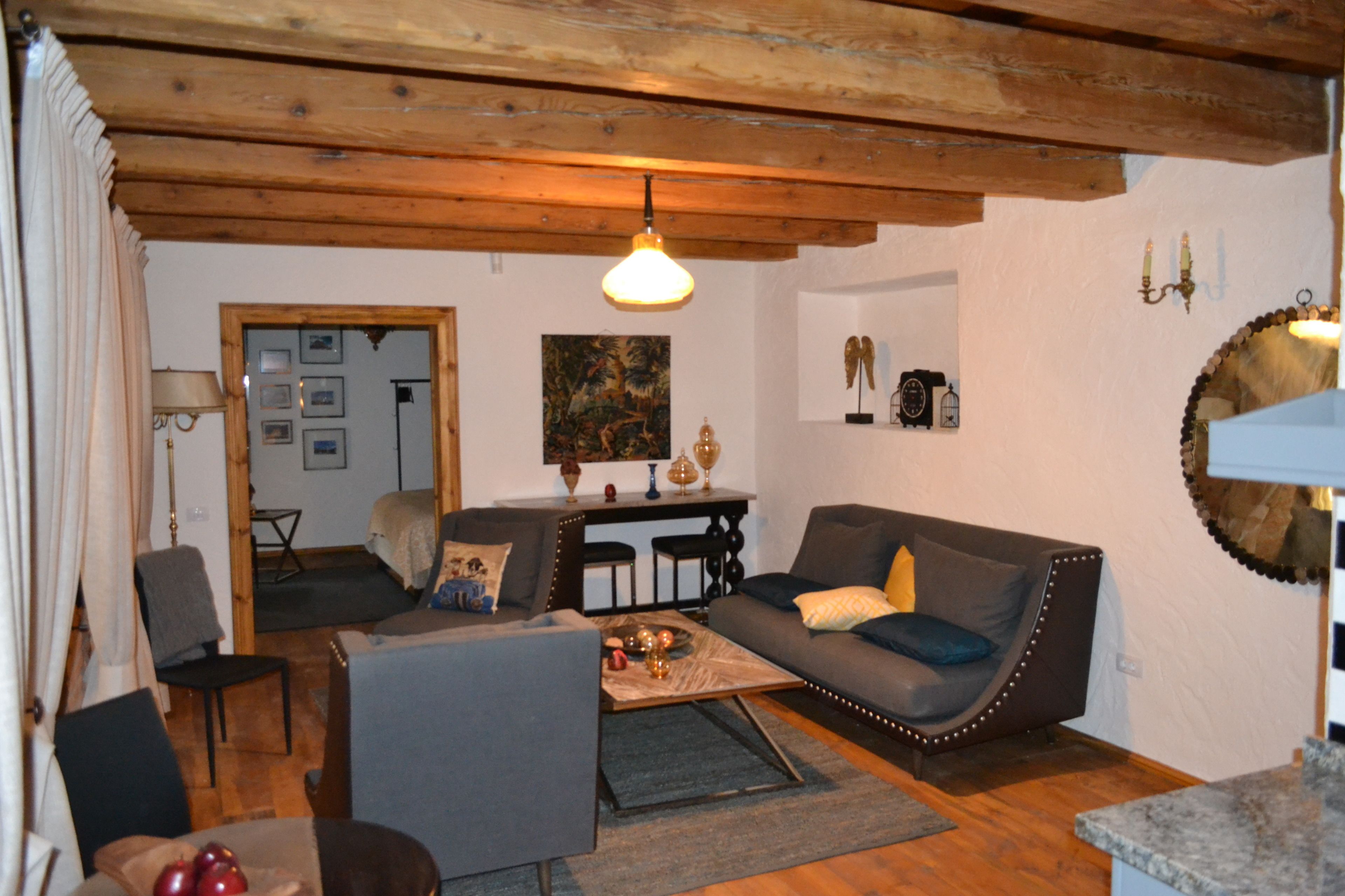 Een gezellige woonkamer met een bank, fauteuil en salontafel. Er is een houten plafond en een spiegel aan de muur.