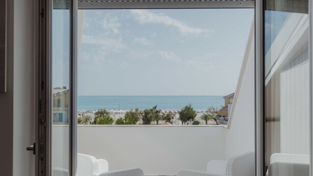 A balcony with white wicker furniture, a clear view of the beach, and a building in the background. - Hotel Caravel (Vasto)