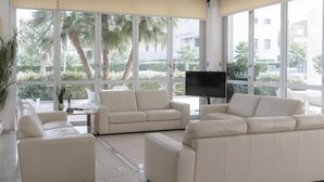 A modern living room with a beige sofa set, a flat-screen TV, and large windows offering a view of palm trees and a building. - Hotel Caravel (Vasto)