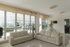 A modern living room with a beige sofa set, large windows, and a potted plant. - Hotel Caravel (Vasto)