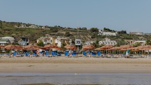 A beach with rows of blue beach chairs and orange umbrellas, a speed limit sign, and houses in the background. - Hotel Caravel (Vasto)