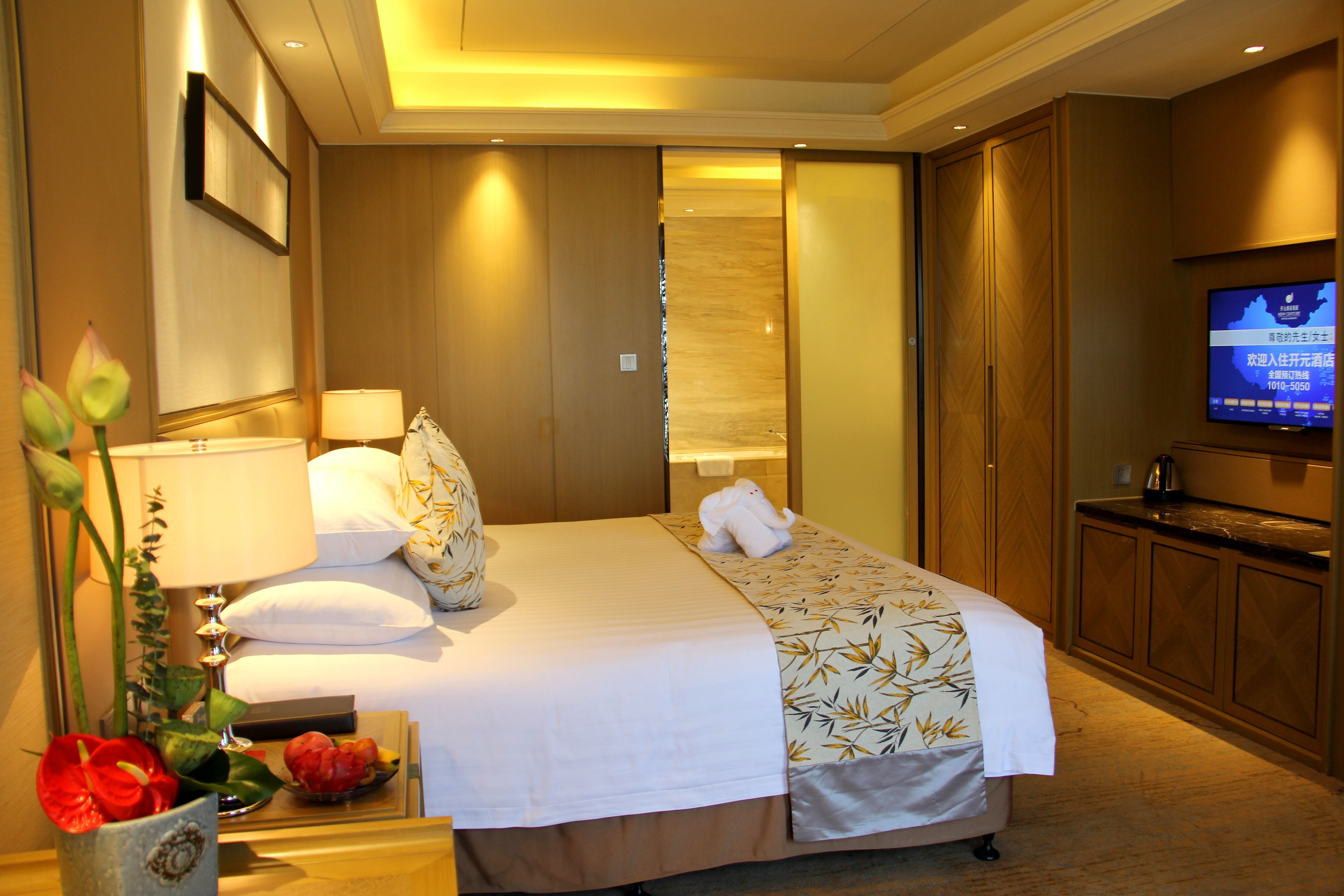 Une chambre d’hôtel avec un lit, des lampes de chevet, une télévision, une armoire en bois et une petite table sur laquelle est posé un vase de fleurs.