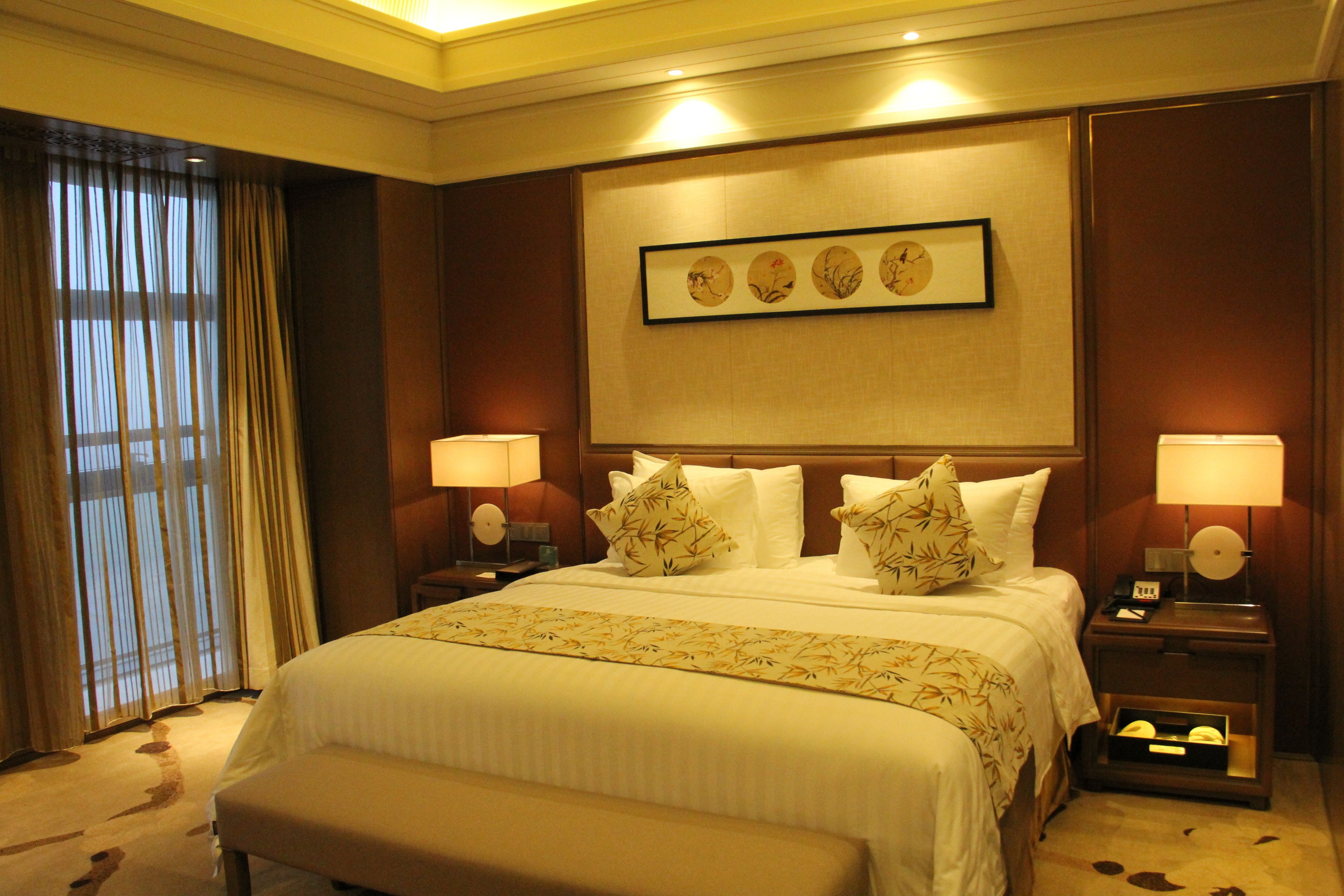 A hotel room with a large bed, two bedside tables with lamps, a headboard with a framed picture, and a window with curtains.