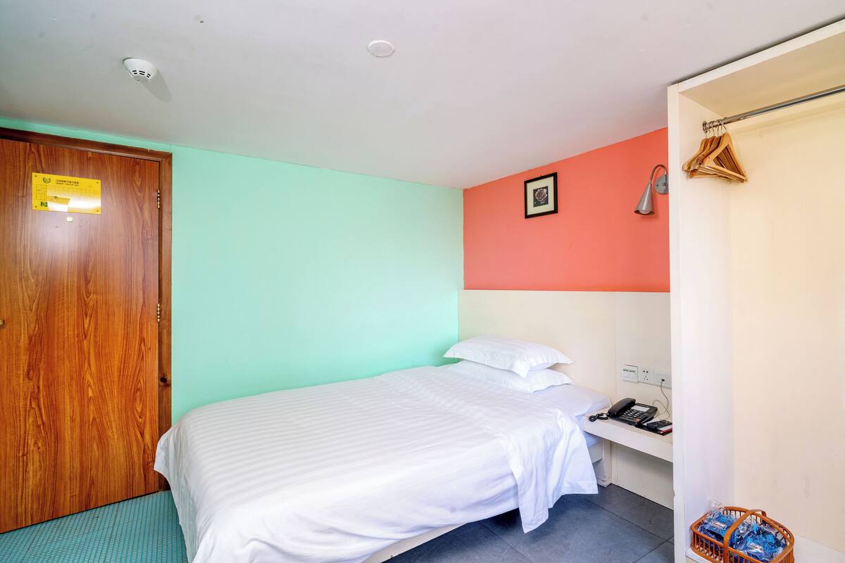 Une chambre avec un lit, une porte en bois, un téléphone fixé au mur et un panier contenant des articles de toilette.