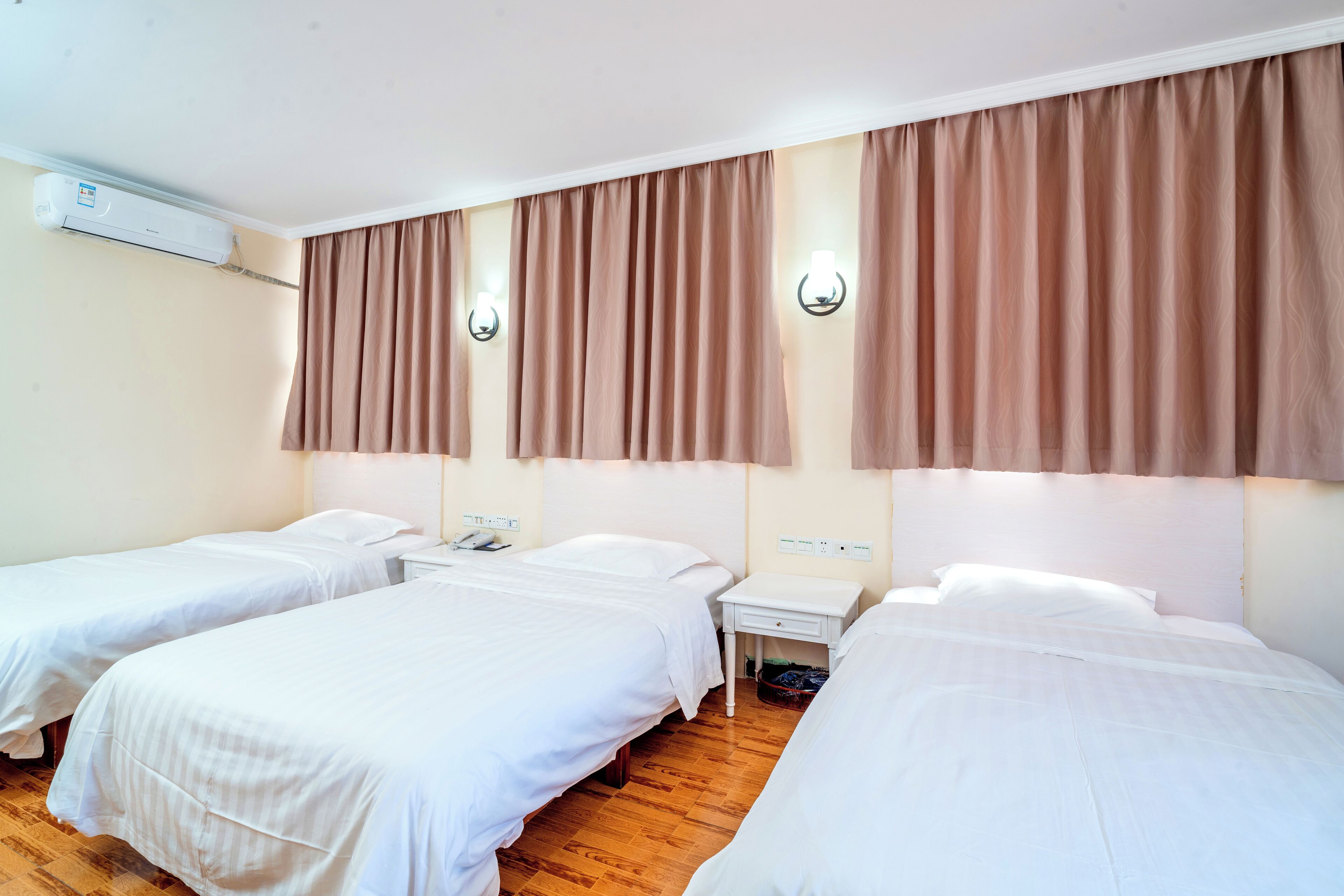 A hotel room with three single beds, a wooden floor, an air conditioning unit, and two wall-mounted light fixtures.