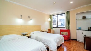 Ein Hotelzimmer mit zwei Betten, einem kleinen Schreibtisch, einem roten Sessel, einer Küchenzeile und einem Fenster mit Blick auf Gebäude.