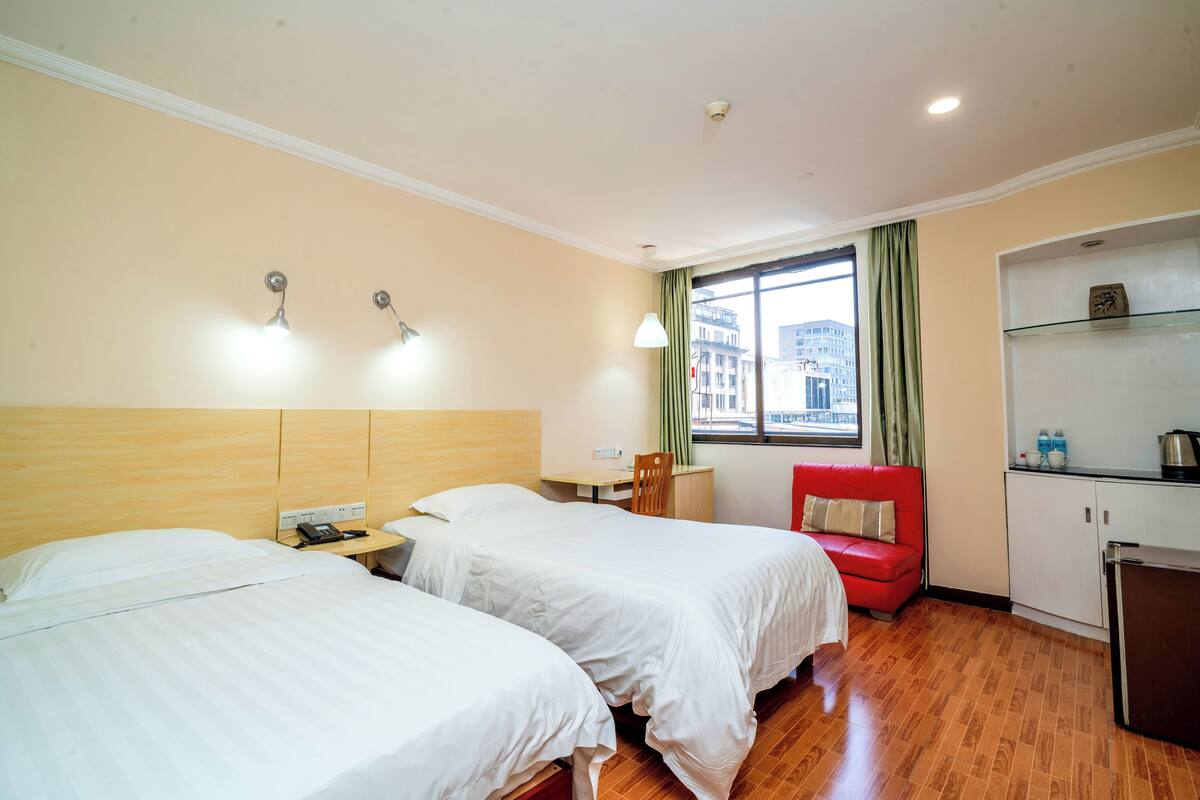 Une chambre d’hôtel avec deux lits, un petit bureau, une chaise rouge, une kitchenette et une fenêtre donnant sur des immeubles.