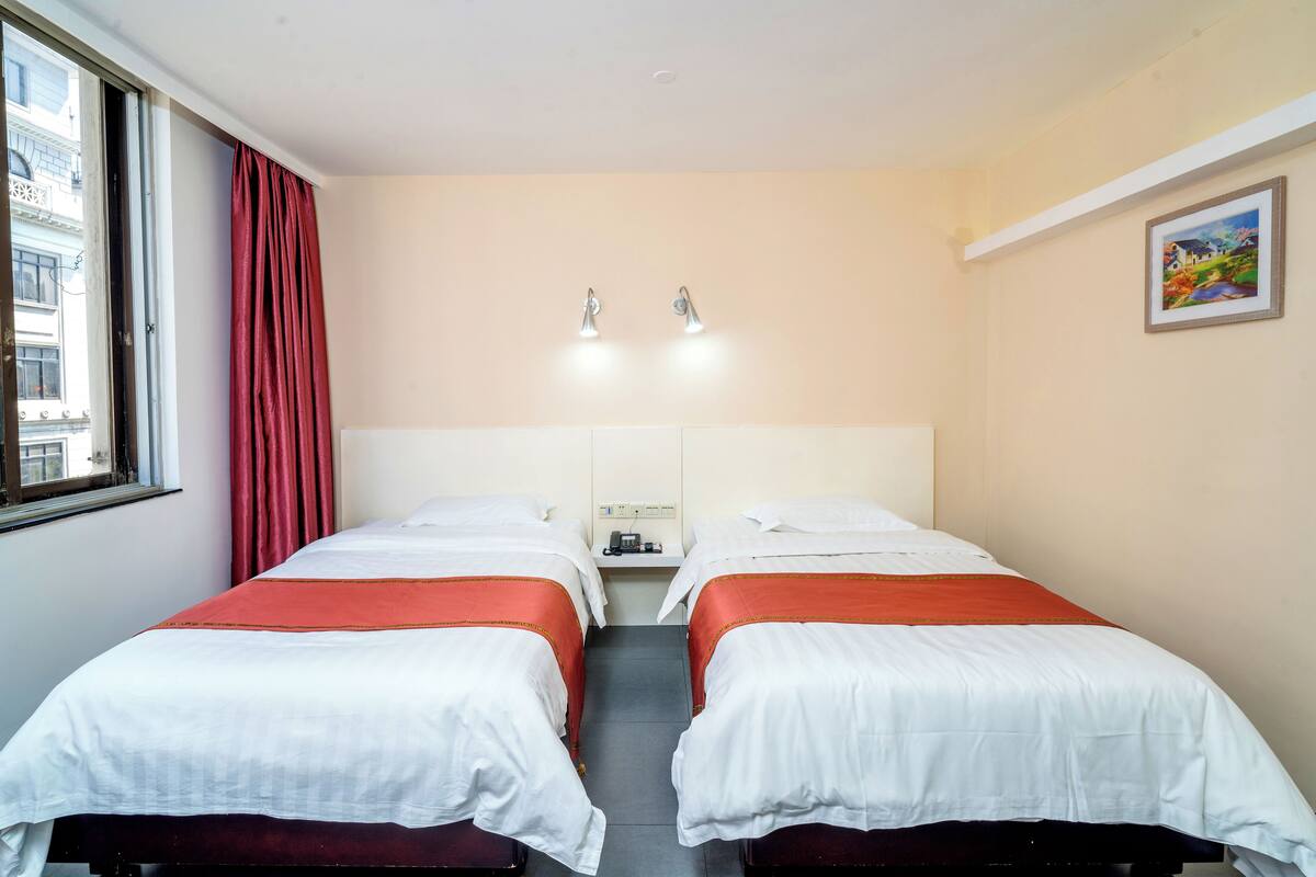 Une chambre d’hôtel avec deux lits, une fenêtre avec des rideaux rouges, un tableau au mur et une table de chevet avec une lampe.