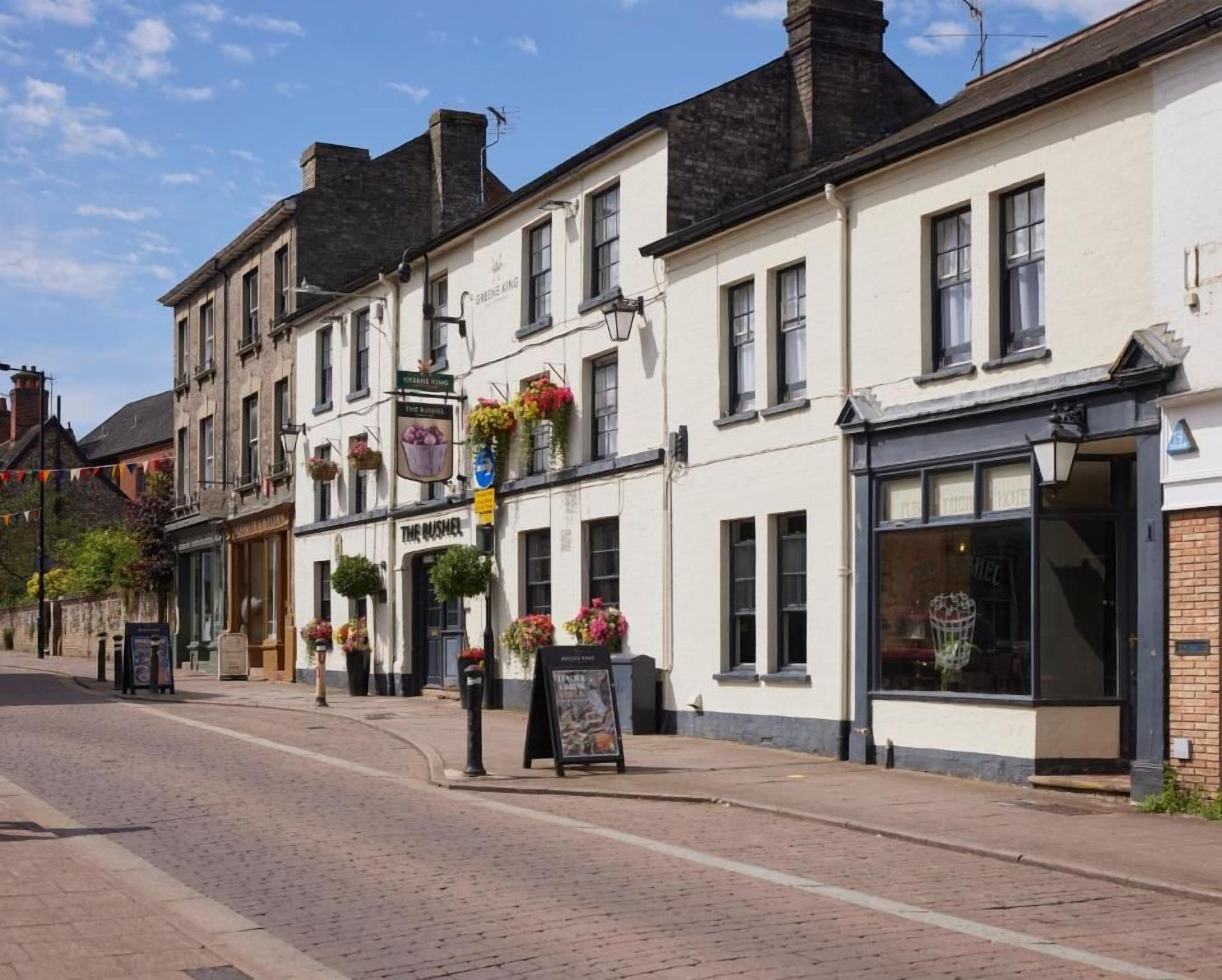 A street with buildings, including a pub with a sign 'The Brusher' and flower baskets on the windowsills.