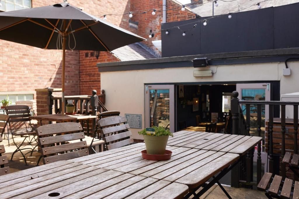 An outdoor seating area with wooden tables and chairs, a potted plant, and a brick building in the background.