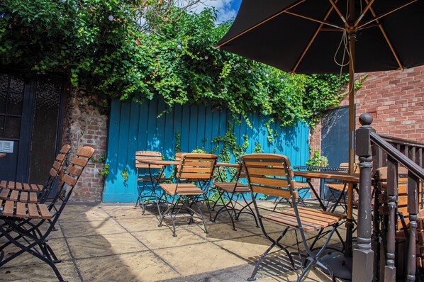 Une terrasse extérieure avec des tables et des chaises en bois, un parasol noir et une clôture bleue recouverte de verdure.