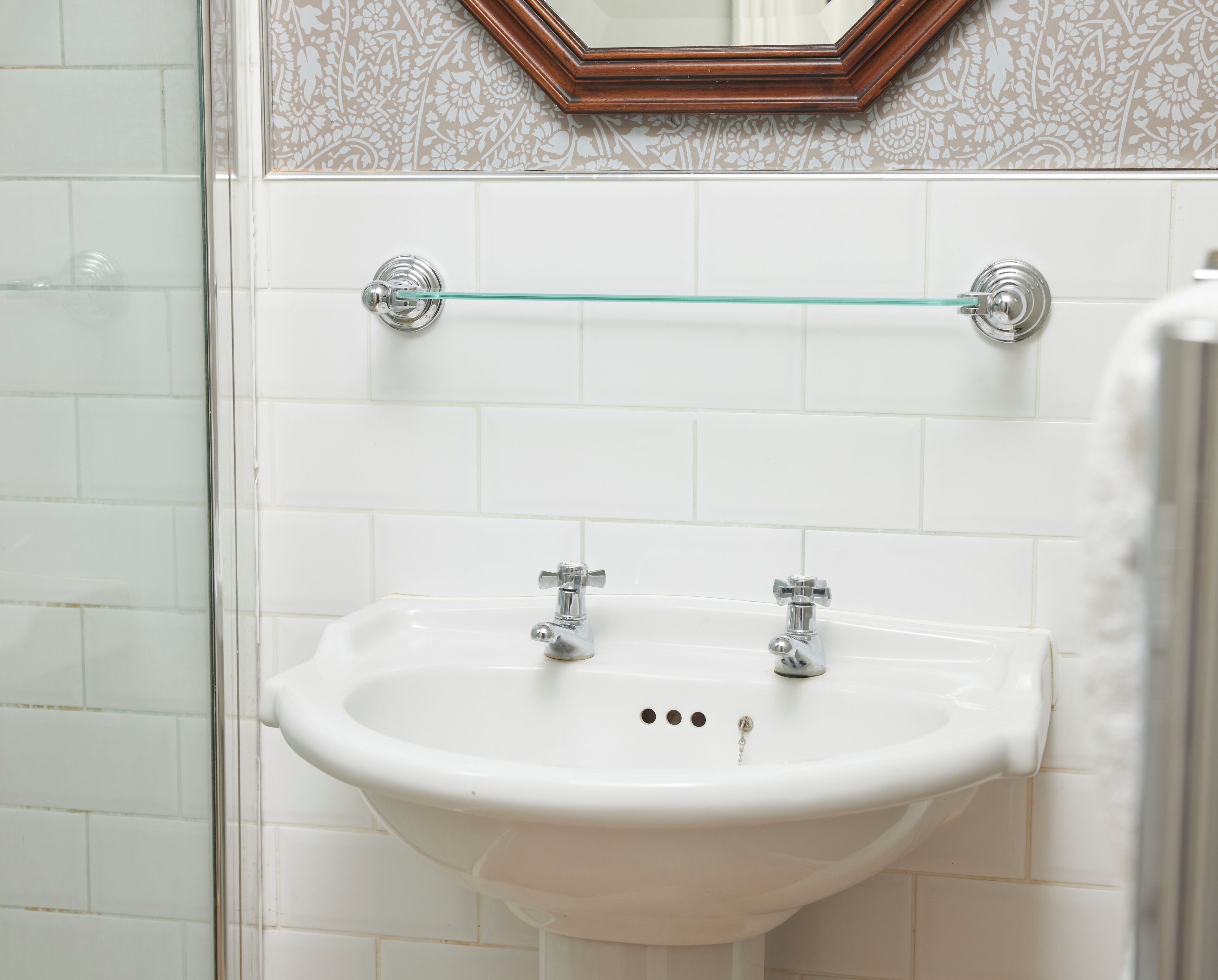 A bathroom with a white sink, two faucets, and a glass shelf above the sink.