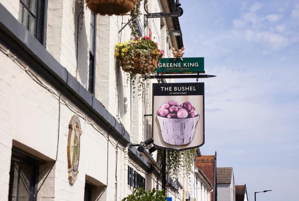 A street sign for 'The Bushel' and 'Greene King' on a building facade.