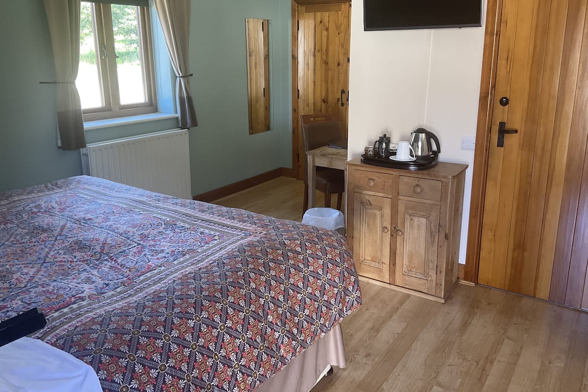A bedroom with a large bed, wooden wardrobe, a small table with a kettle, and a window with curtains.