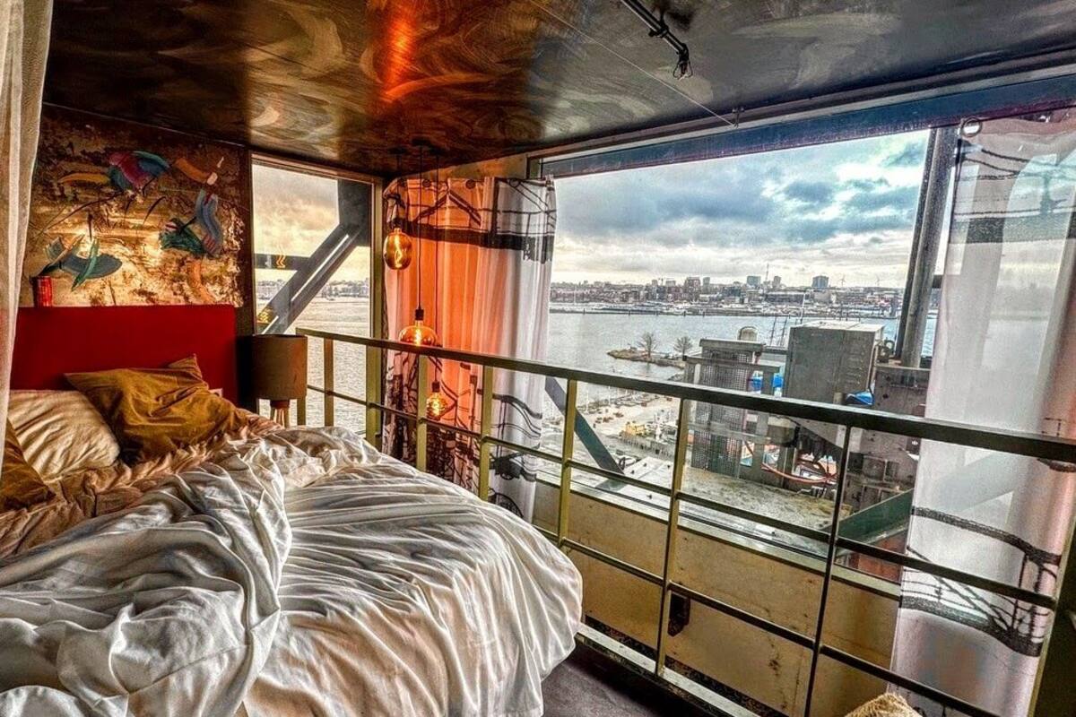 Een slaapkamer met een bed, een stadszicht door grote ramen en een rode muur met kunstwerk.