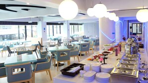 Un espace repas moderne avec des tables et des chaises, un buffet proposant une variété de plats et de boissons, et de grandes fenêtres laissant entrer la lumière naturelle.