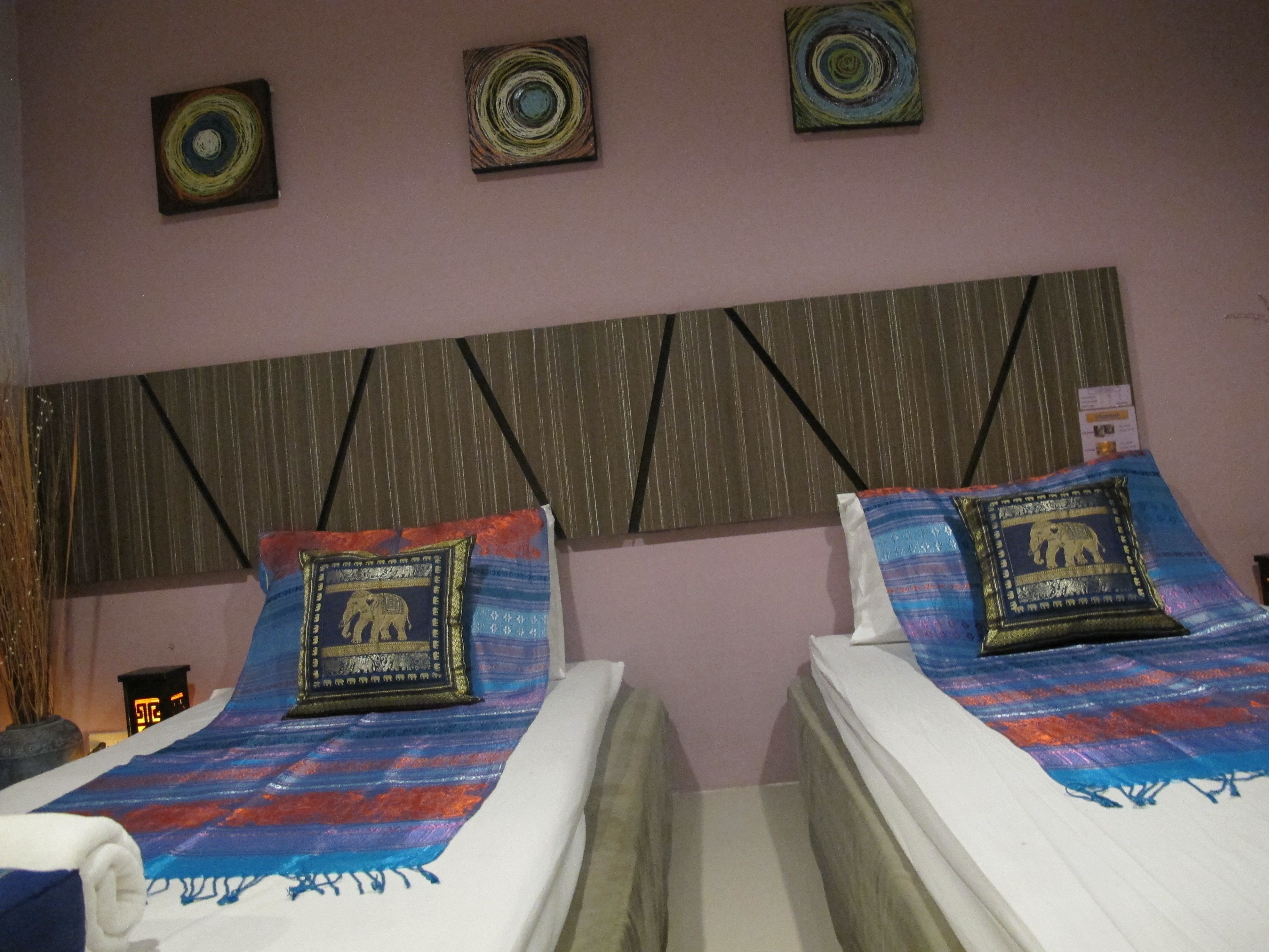 Une chambre d’hôtel avec deux lits simples, chacun agrémenté d’une couvre-lit à motifs et d’un coussin décoratif orné d’un motif d’éléphant. La chambre a un mur rose et trois œuvres d’art murales de forme circulaire au-dessus des lits.