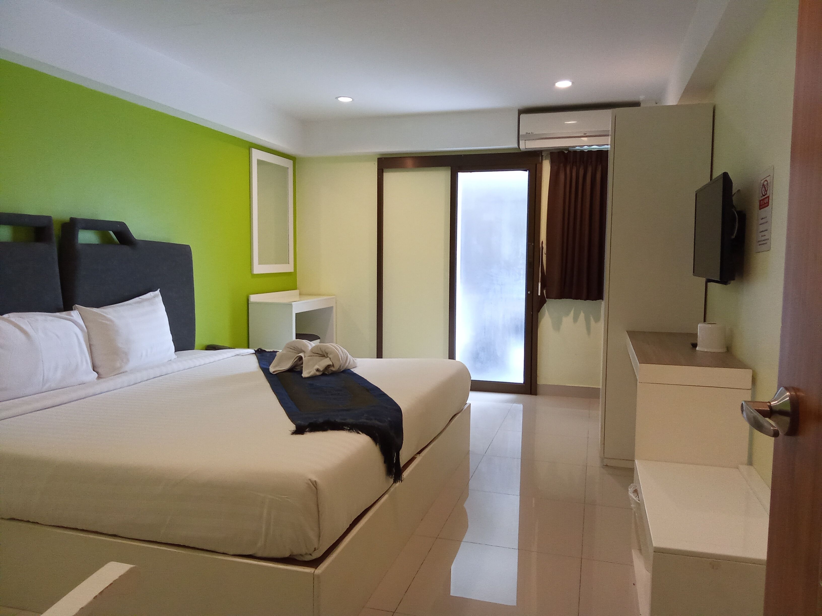 A hotel room with a large bed, a bedside table, a mirror, a TV, and a sliding door leading to a balcony.