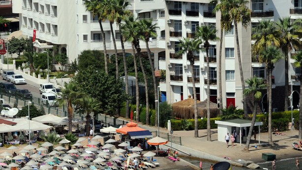 Un hotel costero con varios balcones, una zona de playa con sombrillas y palmeras.