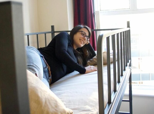 Una persona acostada en una cama dentro de una habitación con ventana y barandilla metálica.
