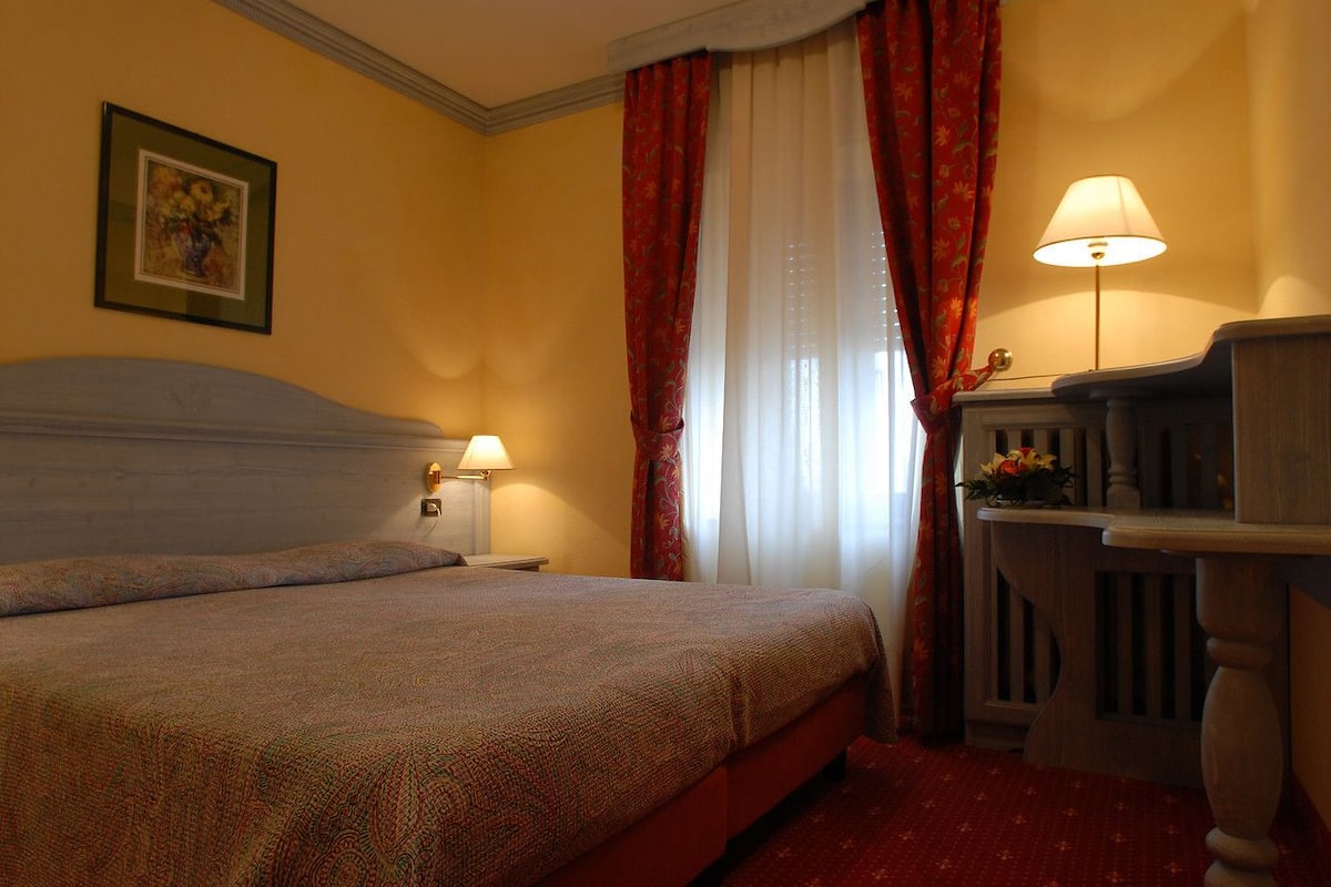 A hotel room with a bed, a nightstand, a lamp, a picture on the wall, and a window with curtains.
