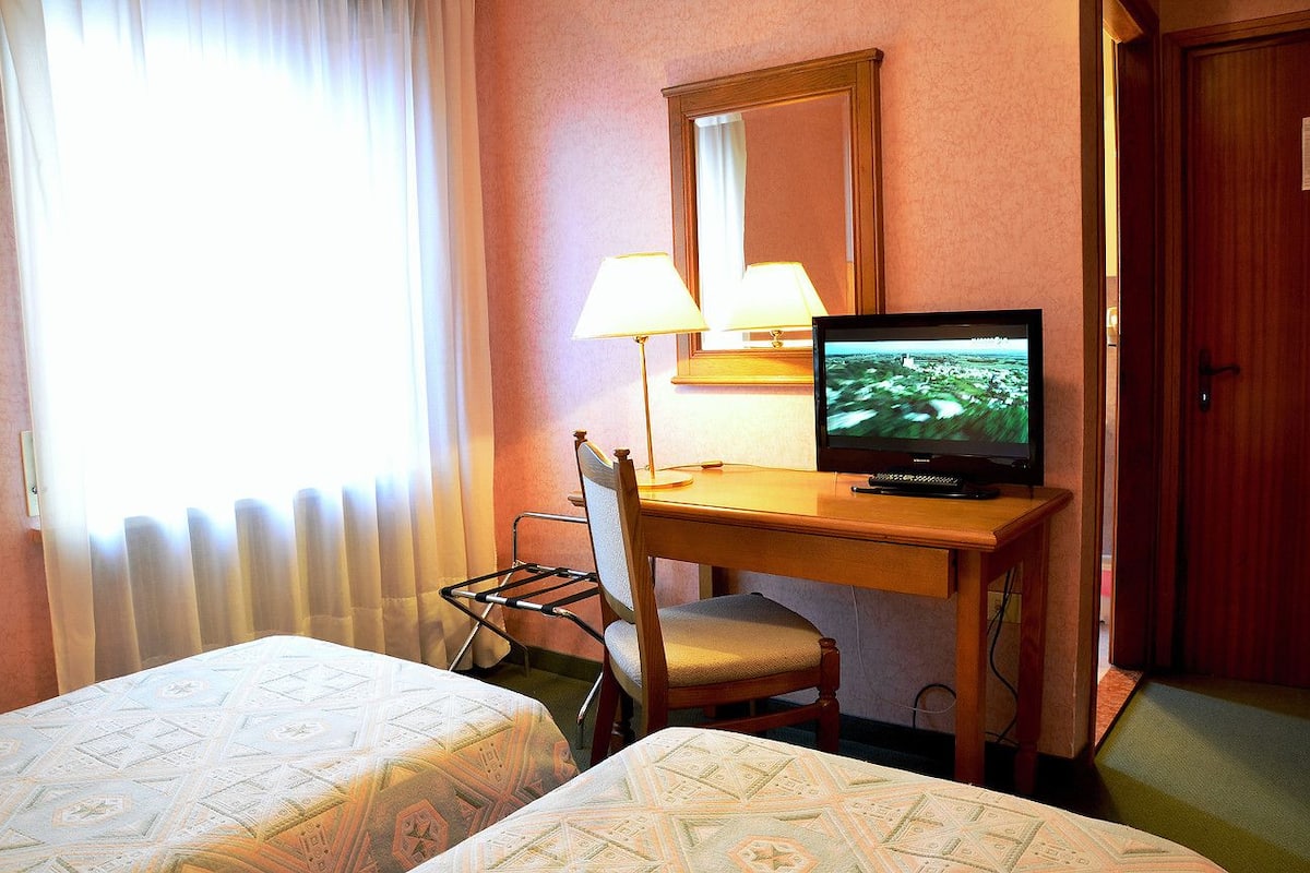 A hotel room with a bed, a desk with a television, a chair, and a window with curtains.