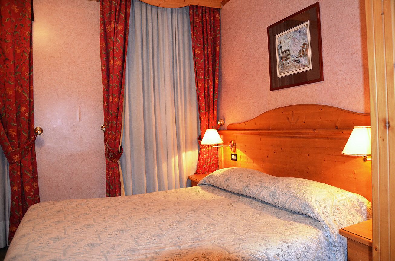 Un dormitorio con cabecera de madera, dos lámparas de noche, cama con ropa de cama estampada, cortinas rojas con diseño floral y un cuadro enmarcado en la pared.