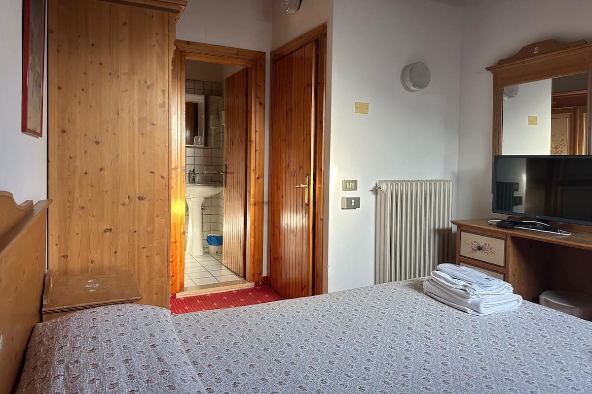 A bedroom with a wooden wardrobe, a bed with a floral bedspread, a radiator, a television, and a mirror on the wall.