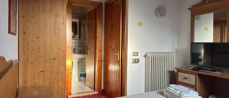 A bedroom with a wooden wardrobe, a bed with a floral bedspread, a radiator, a television, and a mirror on the wall.