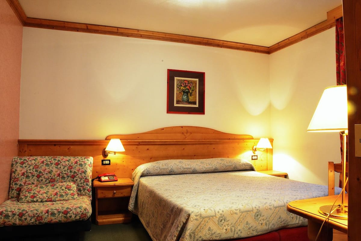 A hotel room with a wooden headboard, two bedside lamps, a floral-patterned chair, and a bed with a patterned bedspread.