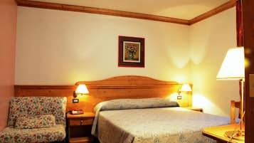 Habitación de hotel con cabecera de madera, dos lámparas de noche, una silla estampada con motivos florales y una cama con colcha estampada.