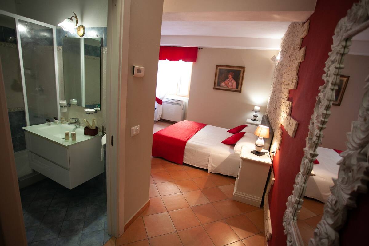 Une chambre d’hôtel avec une salle de bain, un lit recouvert d’une courtepointe rouge, une table de chevet avec une lampe et un tableau accroché au mur.