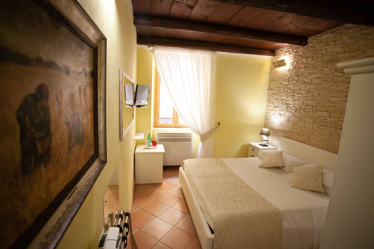 Une chambre à coucher avec un lit, une table de chevet, une lampe, une fenêtre avec des rideaux, un tableau au mur et un mur en pierre.