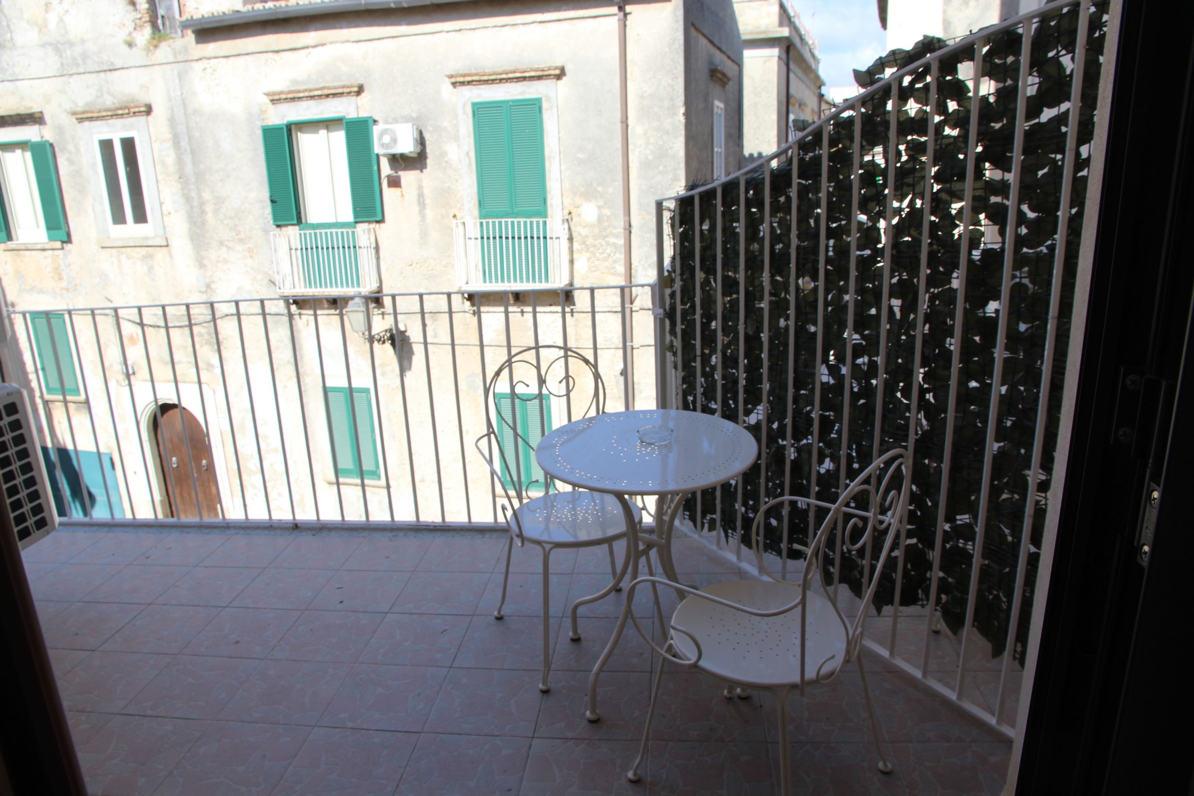 Ein Balkon mit Tisch und Stühlen, einem Metallzaun und einem Gebäude im Hintergrund mit grünen Fensterläden.