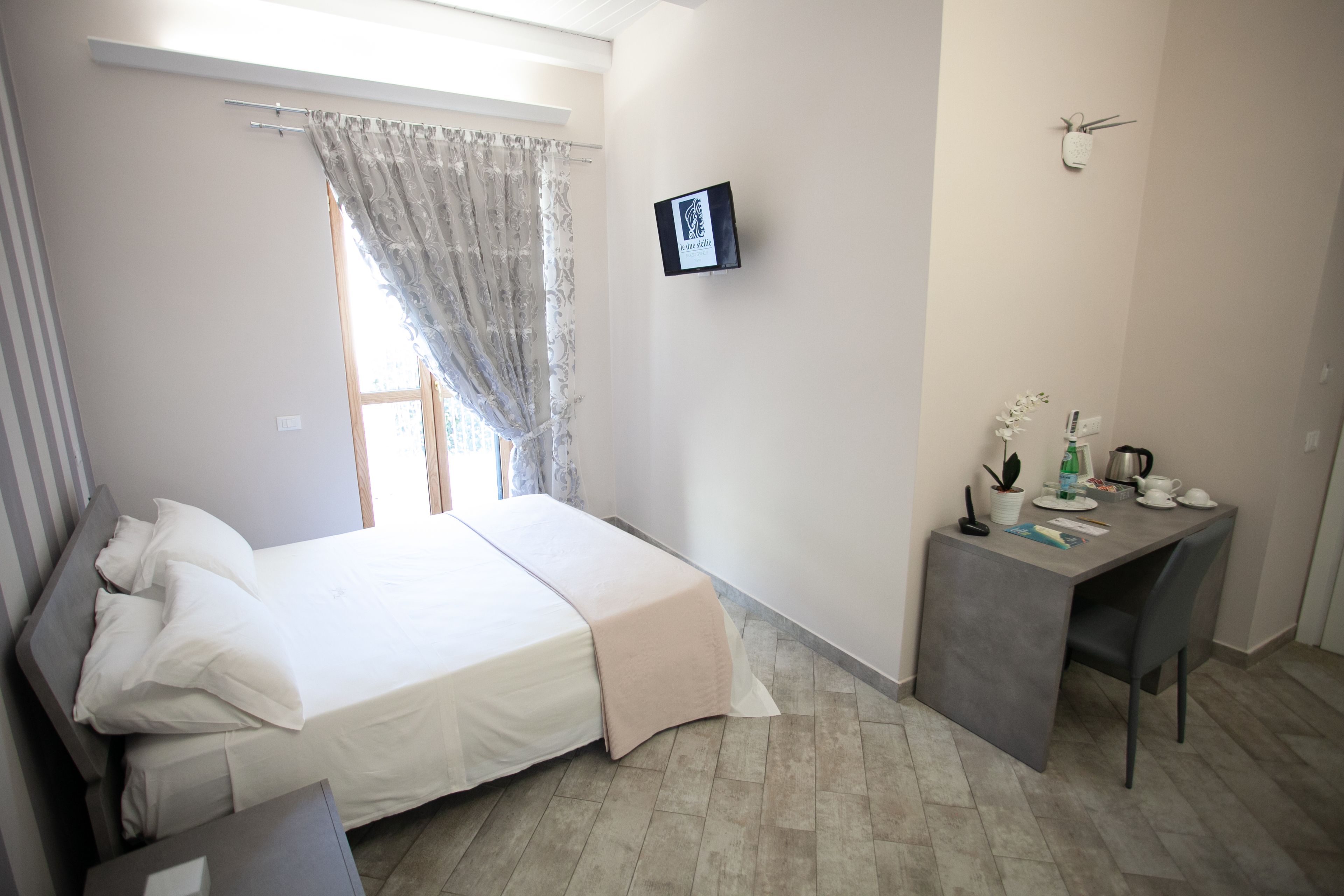 A bedroom with a bed, bedside table, a desk with a chair, a window with curtains, and a wall-mounted picture.