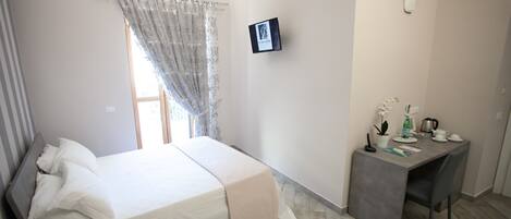 A bedroom with a bed, bedside table, a desk with a chair, a window with curtains, and a wall-mounted picture.