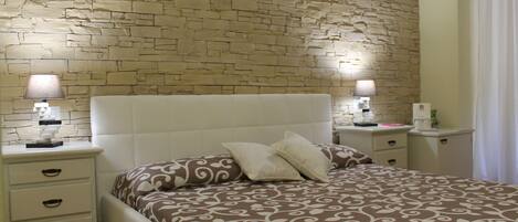 A bedroom with a brick-patterned wall, a white headboard, a patterned bedspread, two bedside tables with lamps, and a nightstand with drawers.