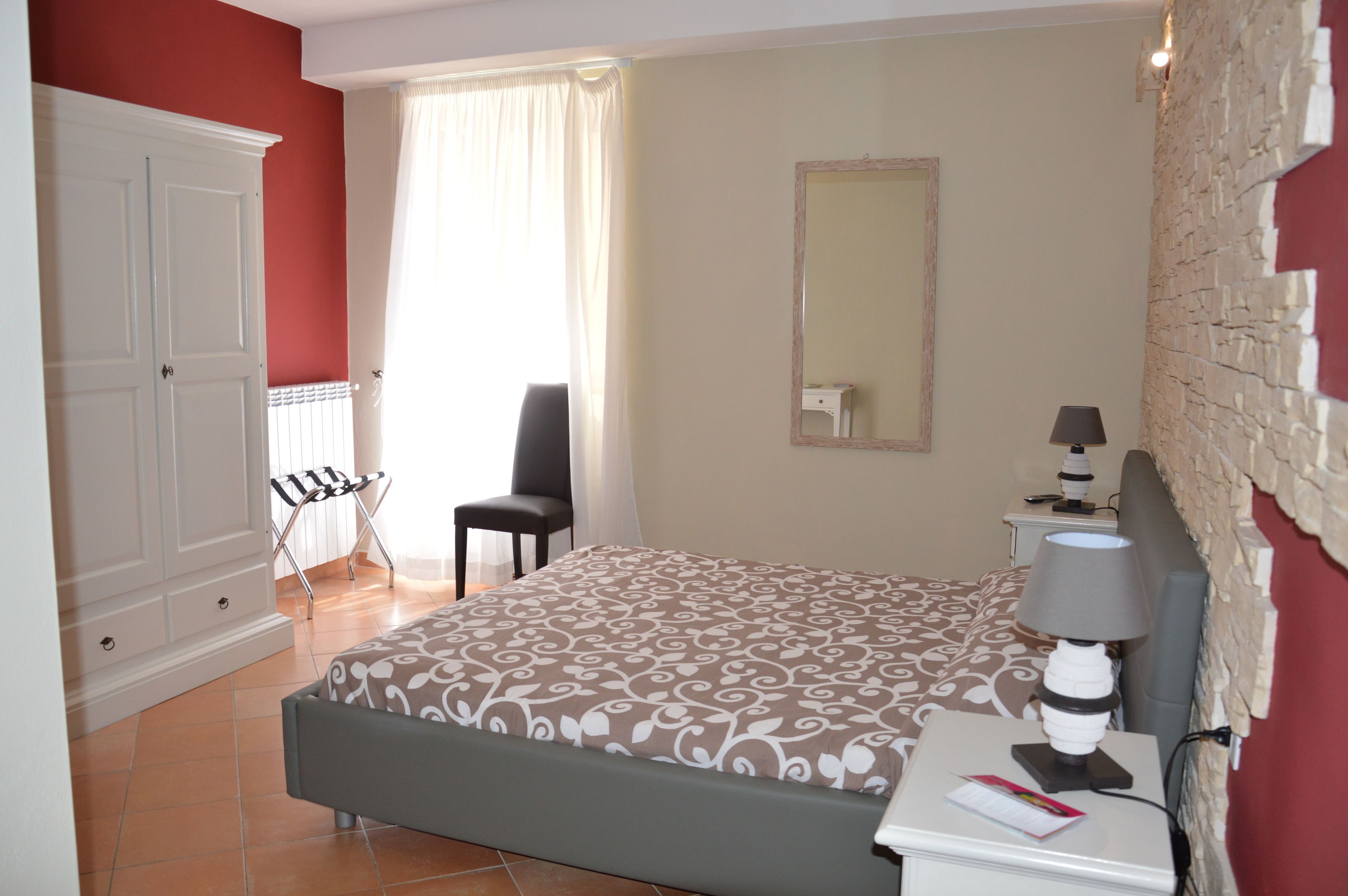 A bedroom with a bed, a chair, a desk with a lamp, a wardrobe, and a mirror on the wall.
