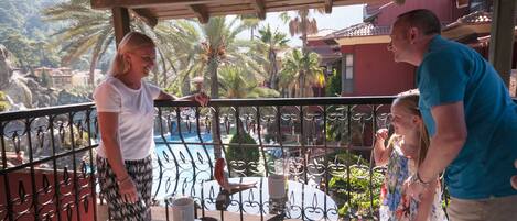 A balcony with a view of a swimming pool, palm trees, and a building with a red facade.