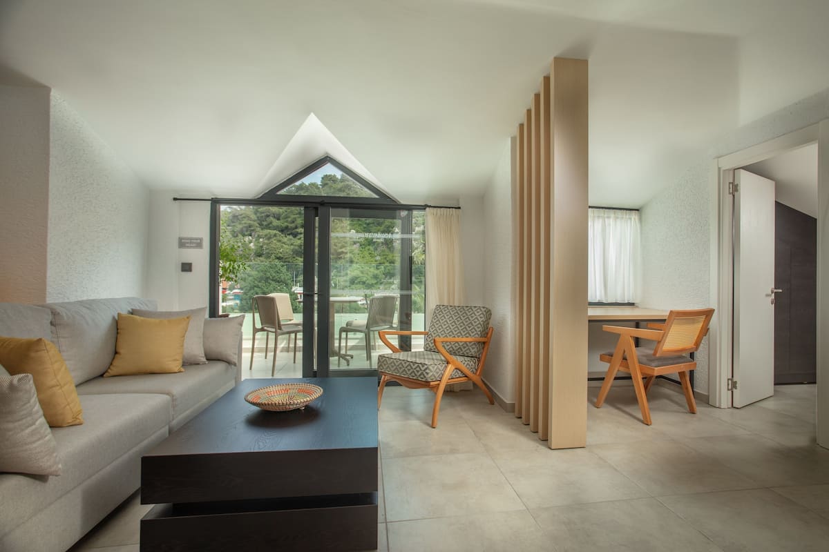 A modern living room with a sofa, coffee table, and chairs. There is a dining area with a table and chairs. A large window provides a view of greenery outside.