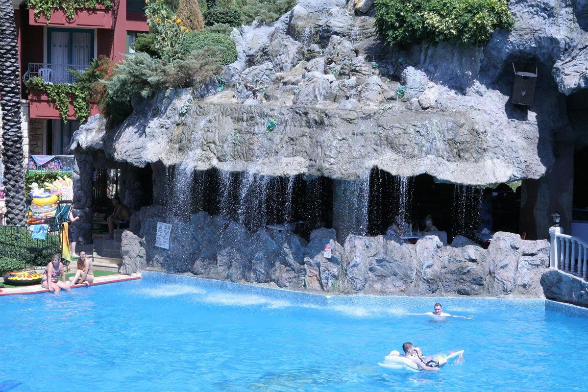 Una piscina con una cascada artificial de rocas y un edificio con balcones al fondo.