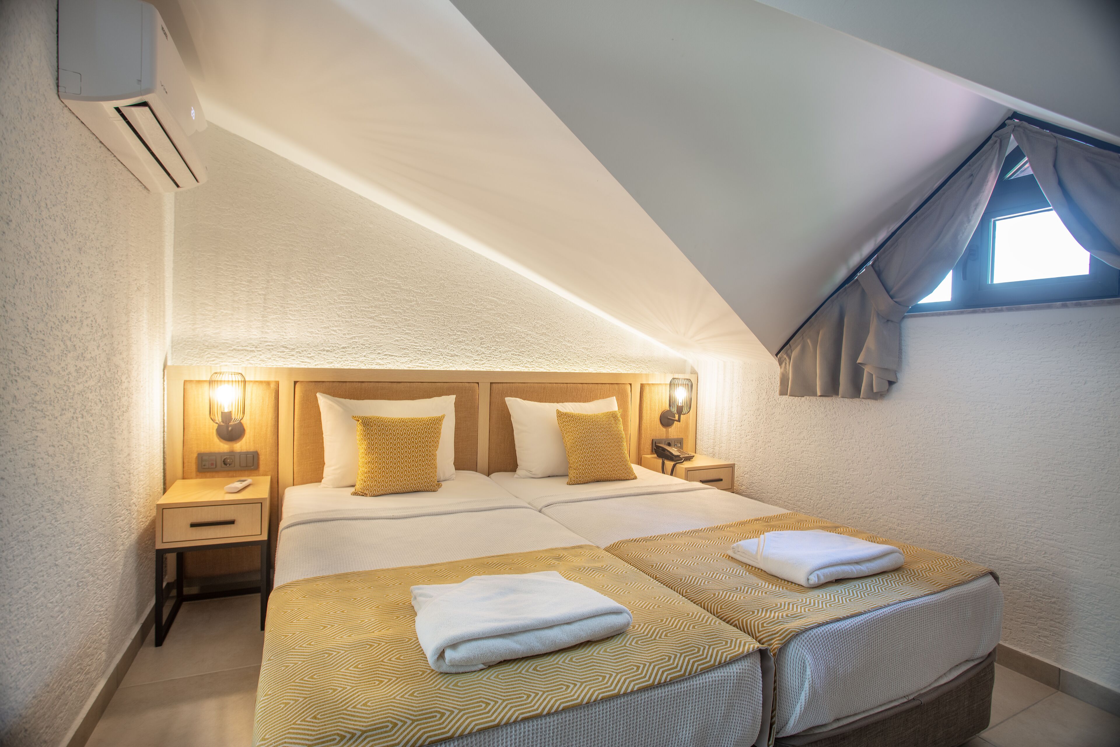 A hotel room with two beds, a nightstand with a lamp, an air conditioning unit, and a window with curtains.