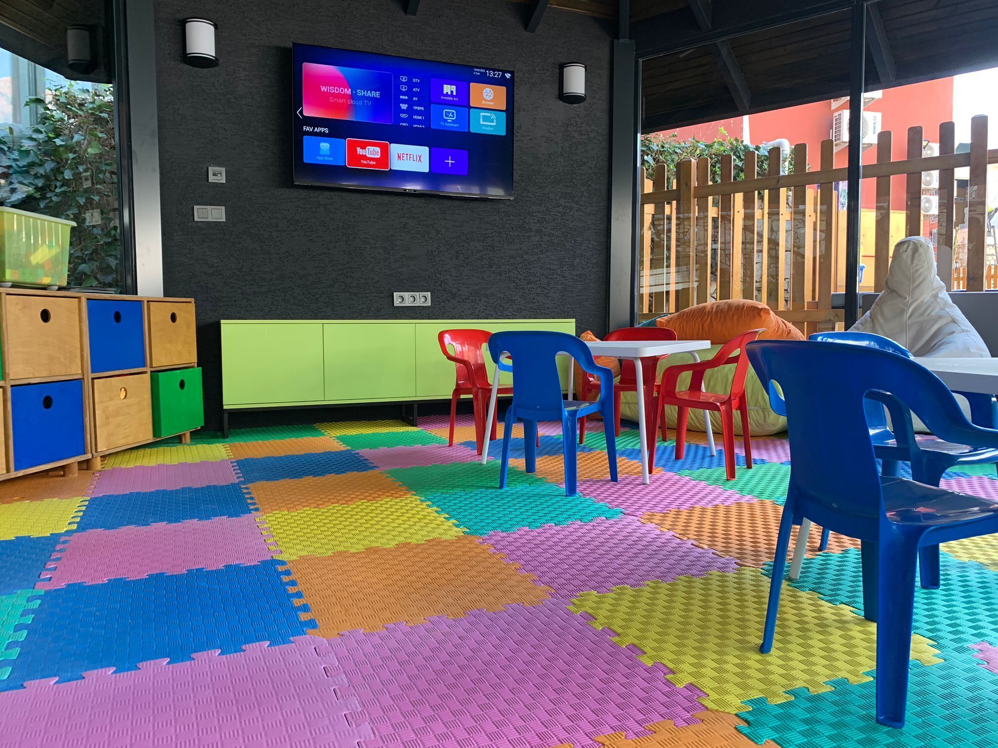 Une pièce dotée d’un téléviseur affichant diverses applications, de tapis colorés et modulaires sur le sol, et d’un espace de jeux en bois avec des boîtes de rangement.