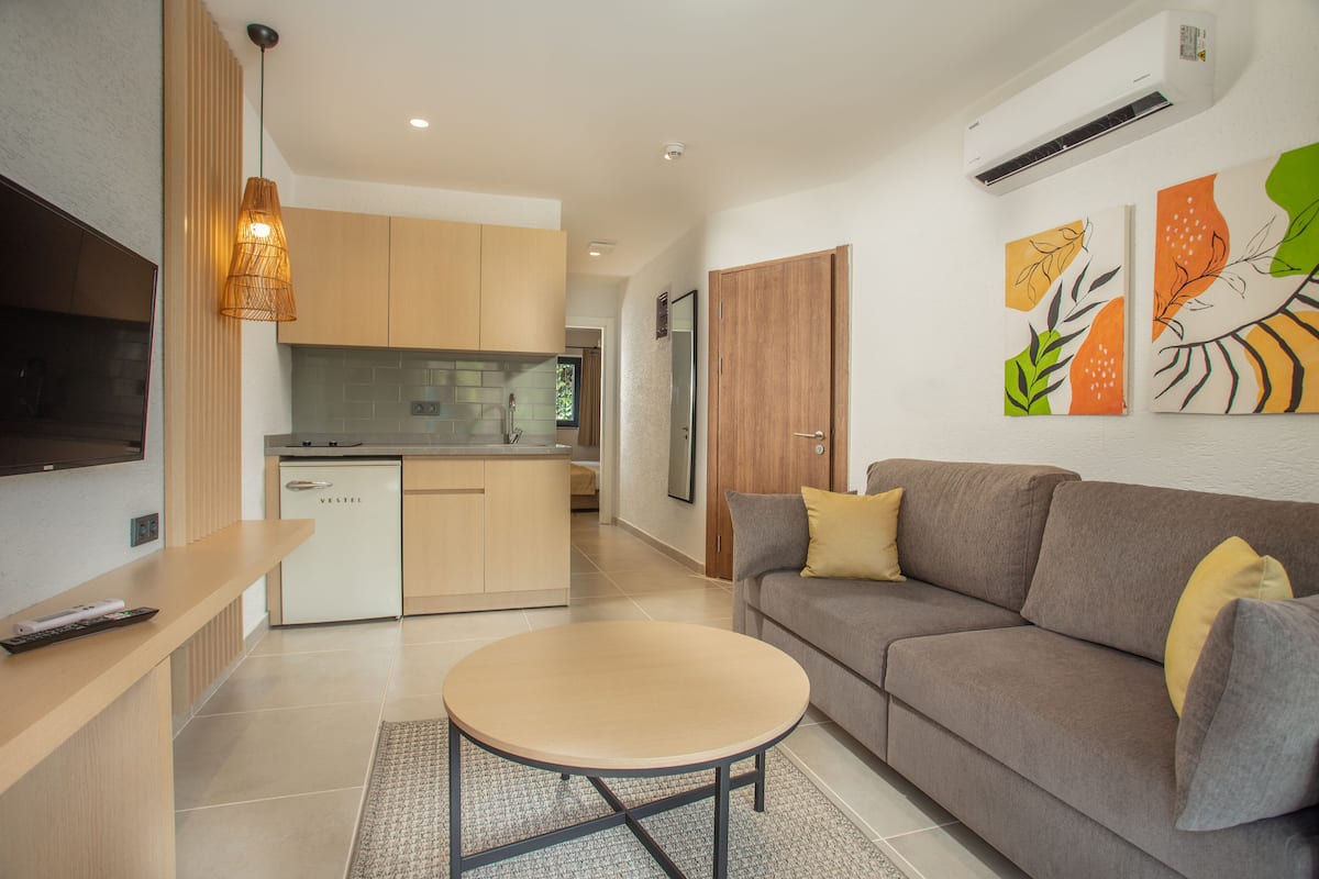 A modern living room with a sofa, a coffee table, and a TV. There is a kitchen area with cabinets and a refrigerator.