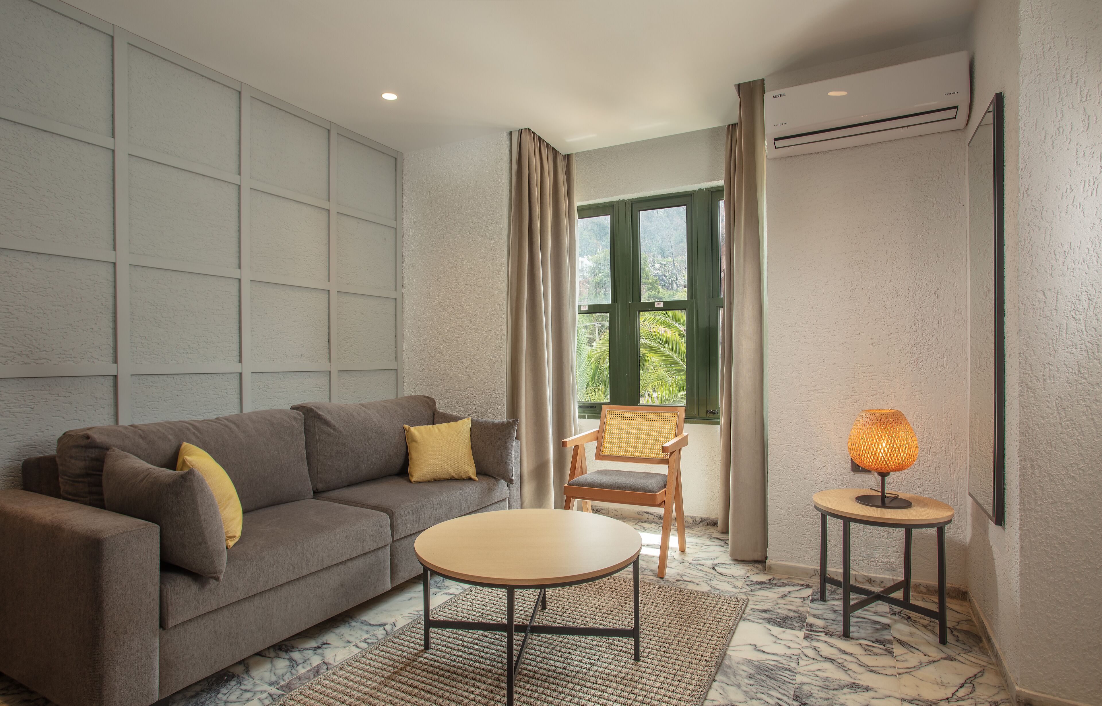 A modern living room with a grey sofa, a round coffee table, a wicker chair, and a small table with a lamp.