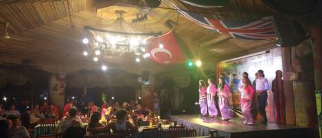 A stage with performers in traditional attire, a crowd of spectators, and a ceiling with hanging lights and flags.