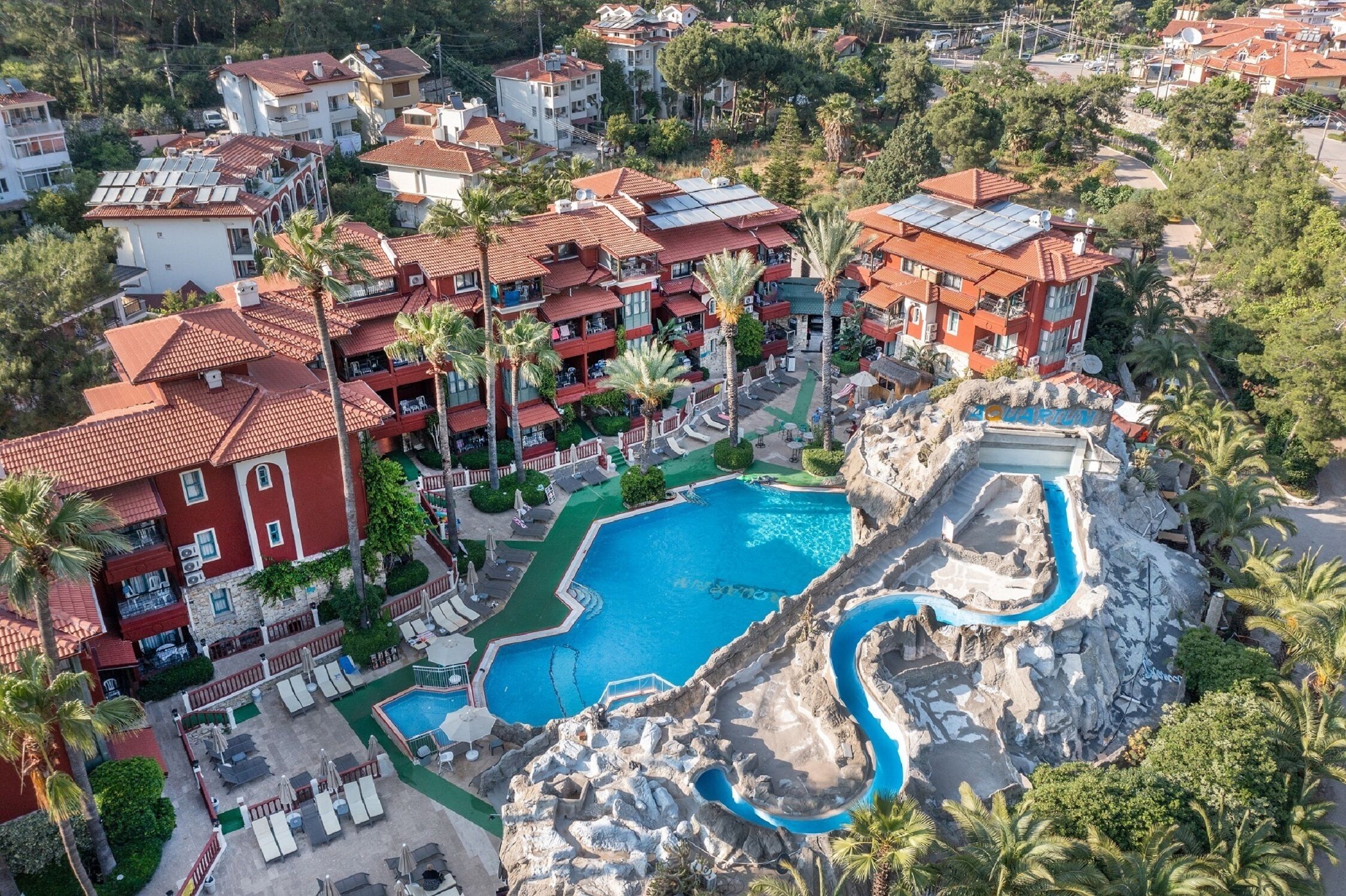 Resort com vários edifícios, piscina central e um elemento decorativo com pedras e água.