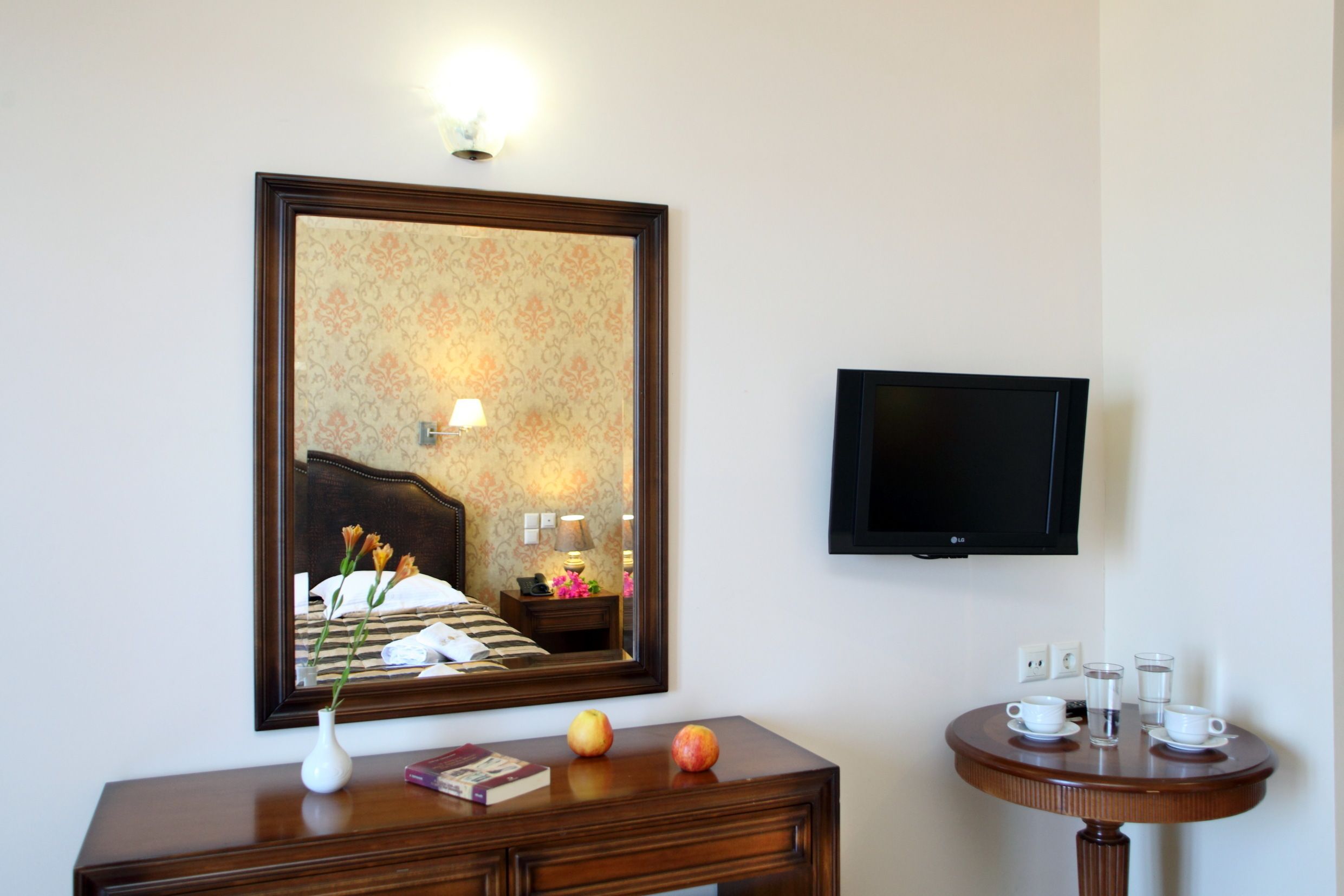 Habitación de hotel con una cama, una mesita de noche con lámpara, un espejo, un televisor montado en la pared y una pequeña mesa redonda con tazas y un florero.