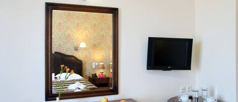 A hotel room with a bed, a bedside table with a lamp, a mirror, a television mounted on the wall, and a small round table with cups and a vase.